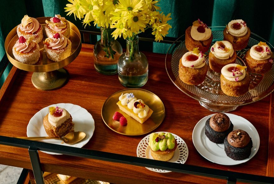 A selection of small cakes and treats on platters, with two vases of yellow daisies.