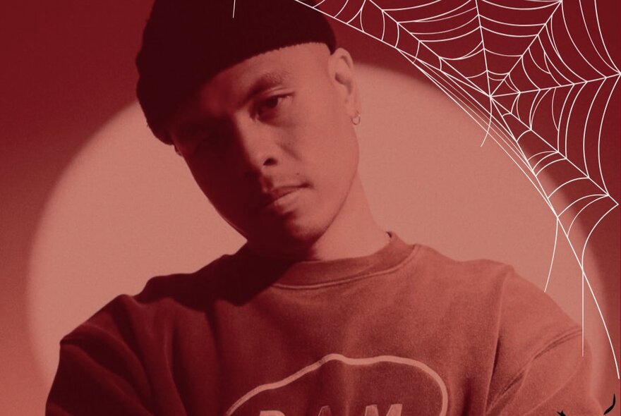 A person wearing a dark beanie and a red sweatshirt with a circular graphic under red lighting, with a white spiderweb pattern in the upper right corner.