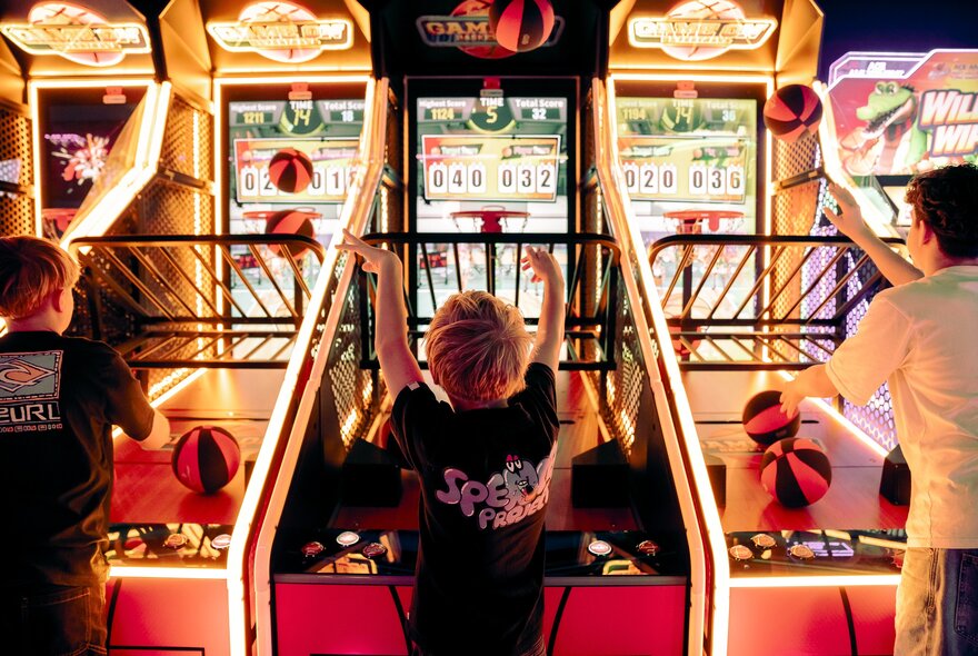 Three teenagers playing a basketball arcade game in an brightly lit games venue.