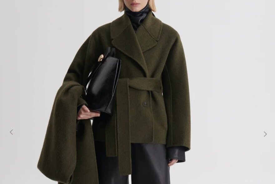 A model is wearing a dark green jacket with a belt and holding a leather bag