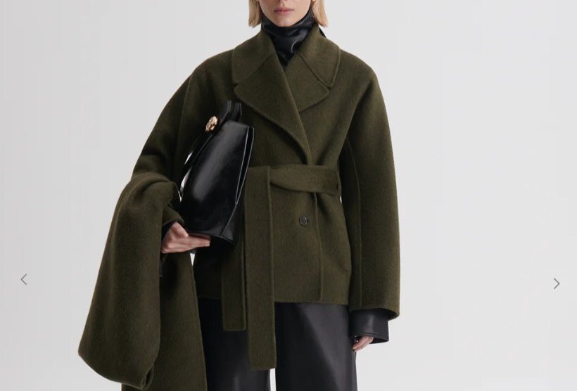 A model is wearing a dark green jacket with a belt and holding a leather bag