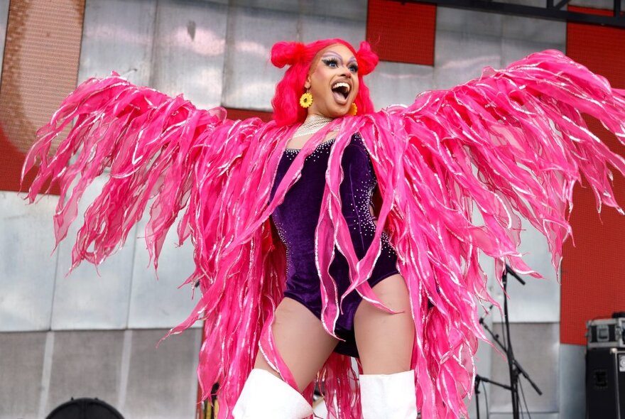 A person wearing pink feather wings with a bright red wig, and thigh high white boots, performing on a stage outdoors. 