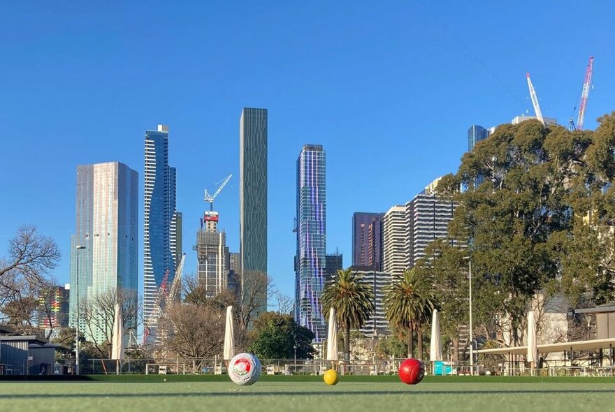 Bowls club at eye level with grass playing area with balls set against city skyline of tall buildings, trees and blue sky.