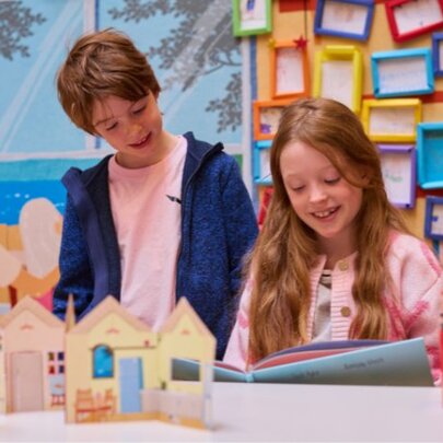Two children stand at a table looking at a craft project in a room that features a colourful display of framed artwork or pictures hung in a grid pattern on the wall behind the table.