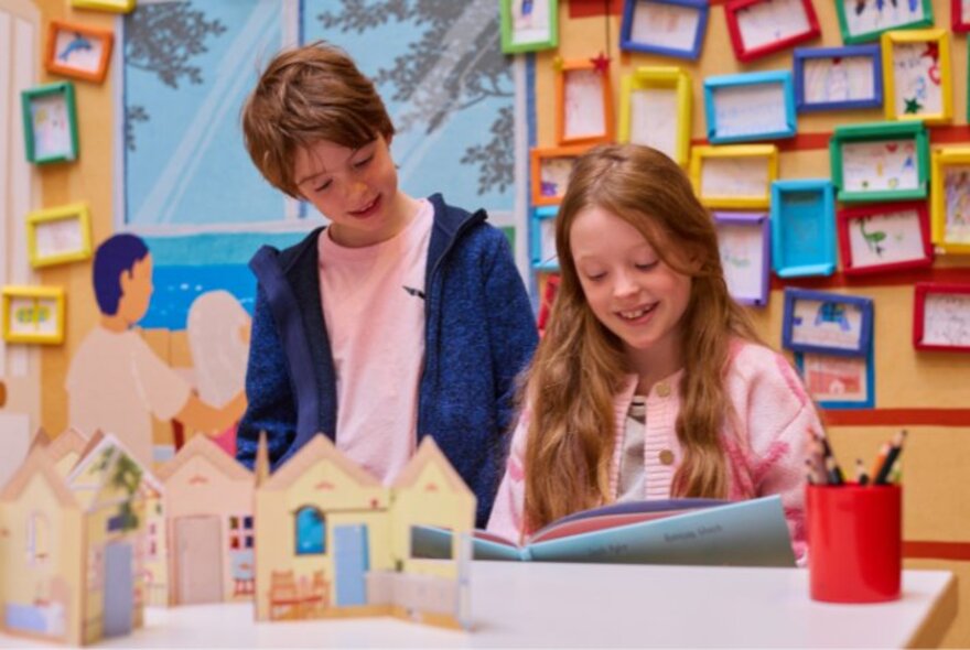 Two children stand at a table looking at a craft project in a room that features a colourful display of framed artwork or pictures hung in a grid pattern on the wall behind the table.