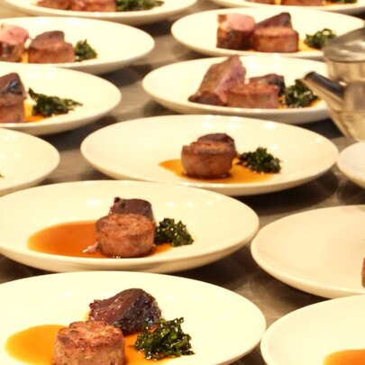 Rows of white plates arranged on a stainless steel countertop in a professional kitchen, each dish contains a thick cut of meat, in an orange sauce, and a small serving of dark green leafy vegetables.
