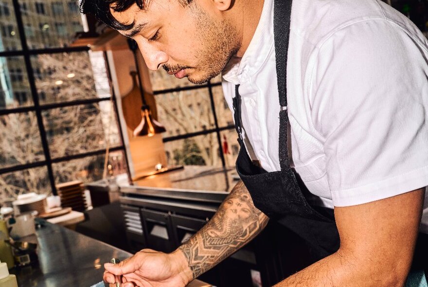 A tattooed chef plating food in a restaurant kitchen.