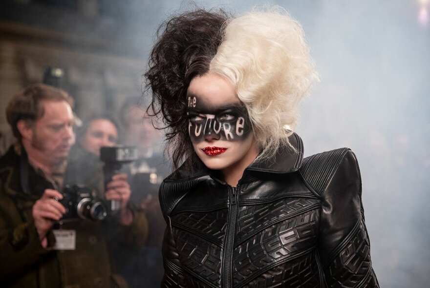 A still from the film Cruella with an actor wearing a black leather jacket, with black face make-up and a teased up hair-do that is half white-blond and half black.