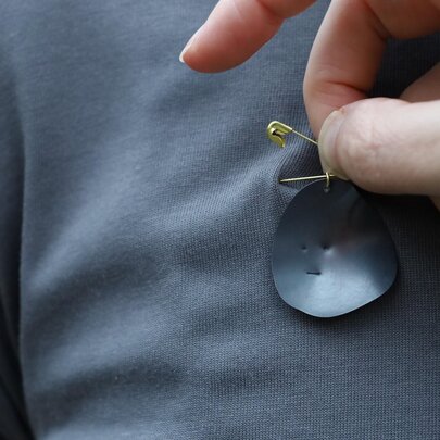 A hand attaching a small, black oval metal disc to a grey t-shirt with a safety pin.