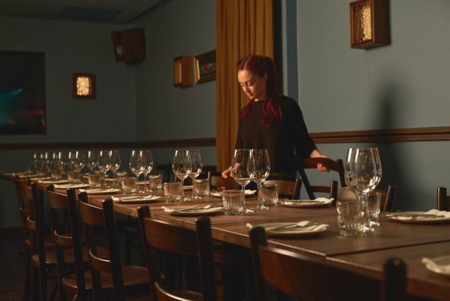 A waitress wearing black, straightening a plate on a dining table with wine glasses and plates for 16 place settings in a low-lit private dining room.