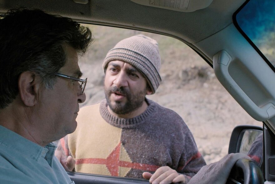 A still from an Iranian movie showing a man in a beanie talking to a driver through the front window of a car.