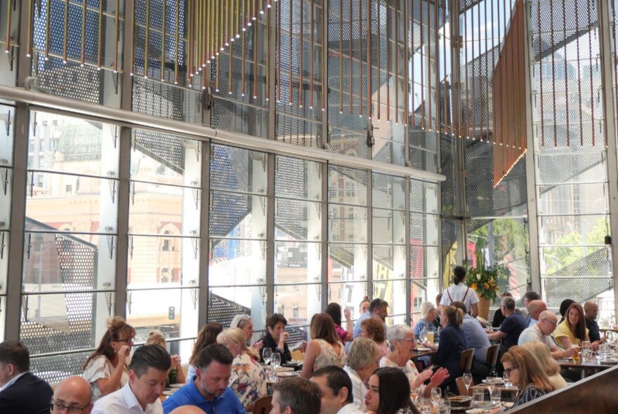 Airy dining space at Taxi Kitchen, with huge windows and overhead lighting, and patrons seated at tables eating and drinking.