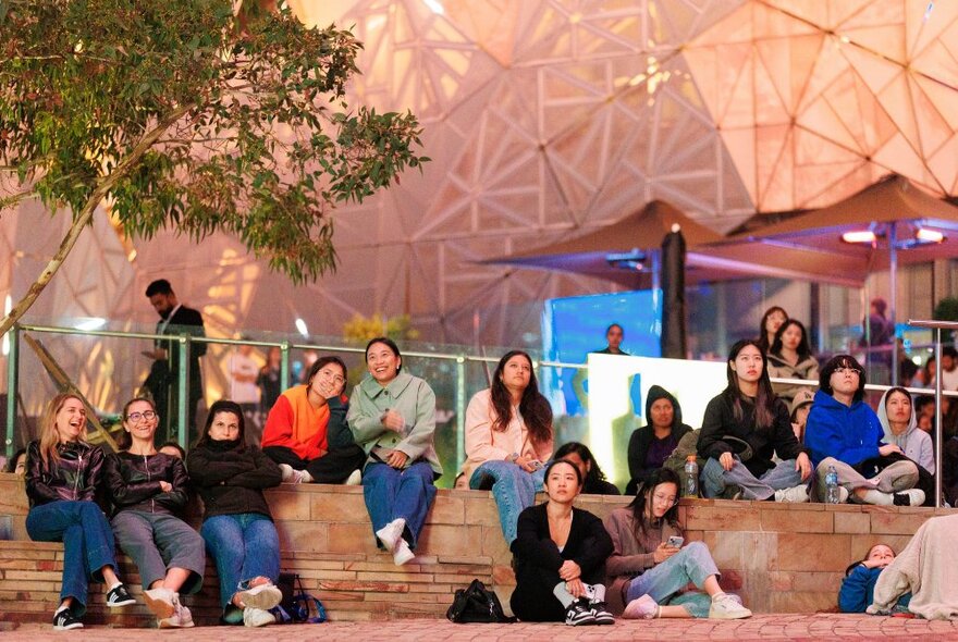 People sitting on the stairs at Fed Square at night, looking up.