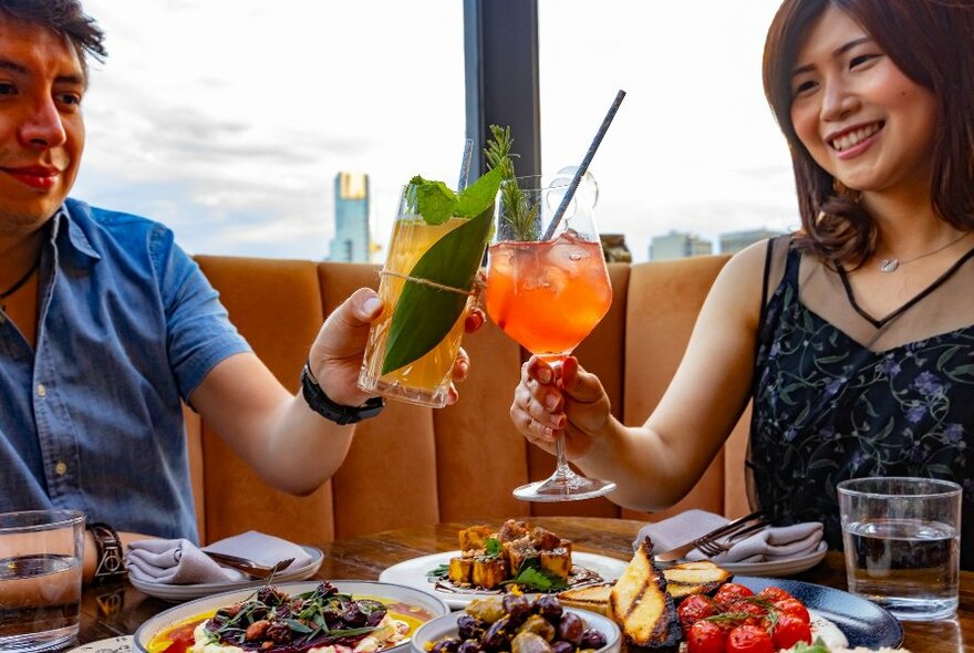 Two people dining on a rooftop clinking two cocktails together over a table full of food.