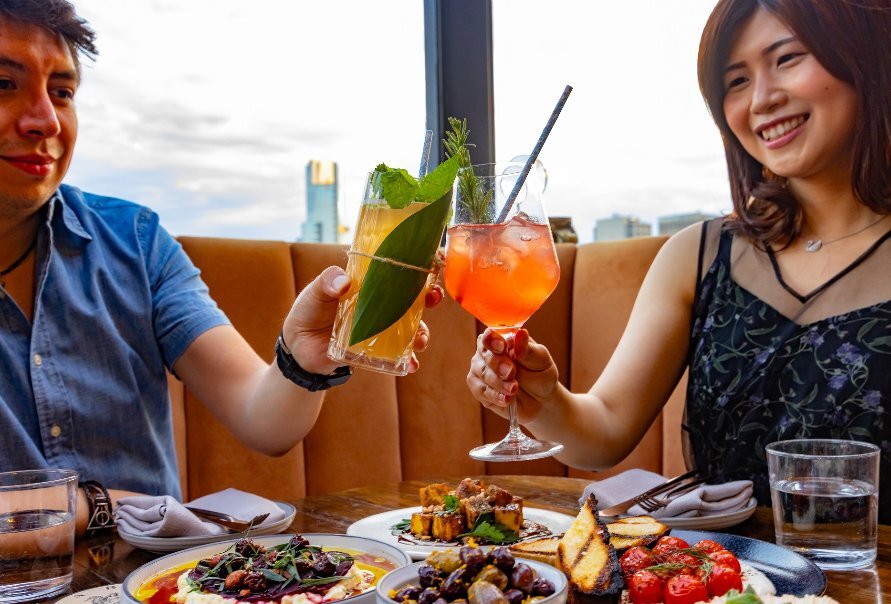 Two people dining on a rooftop clinking two cocktails together over a table full of food.