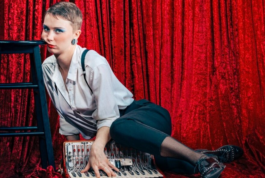 Person seated on a floor of a stage, a red curtain backdrop, the side of their face pressed up to the edge of a stool, and with their hands on a piano accordion that sits on the stage floor.