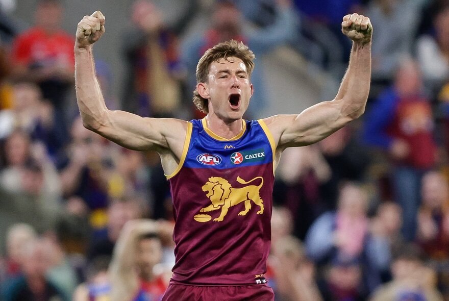 A Brisbane Lions AFL player celebrating on the field with his fists clenched and arms raised above his head, with blurred fans in the stadium in the background.