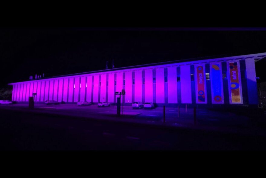 The external facade of a long building lit up purple at night.