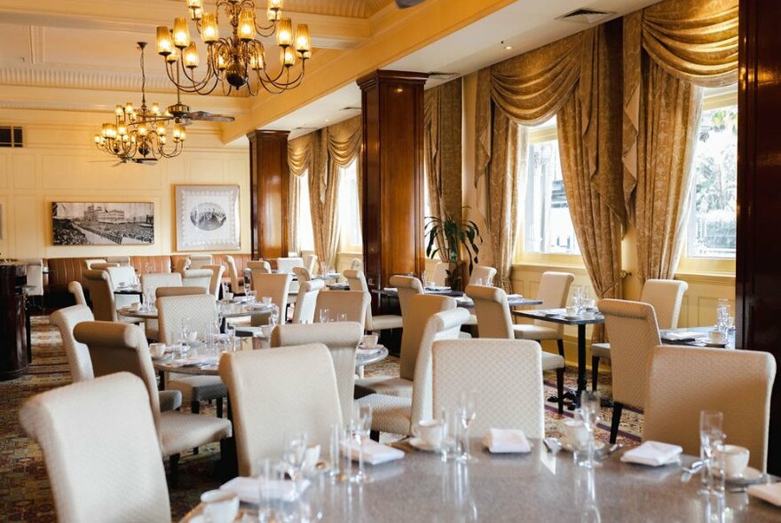 Interior of The Windsor Hotel dining room, with round tables set for service, gold curtains framing large windows, and chandeliers hanging from the ceiling.