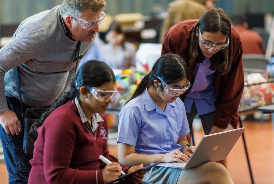 Two young people wearing protective eyewear while they work on personal laptop or tablet devices, watched on by two adults also in protective eyewear.