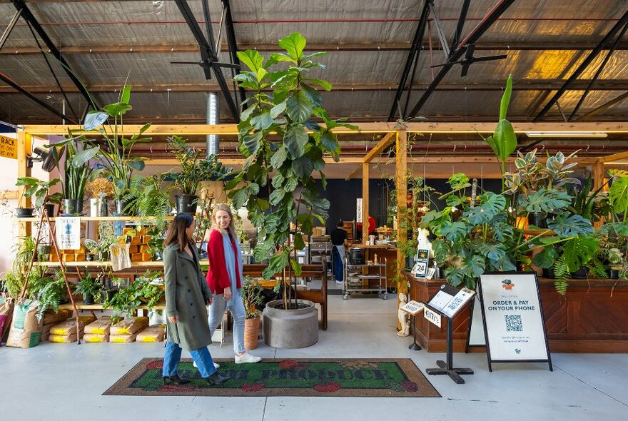 Two women walking into a cafe in a warehouse, with plant-filled shelves dividing the space.