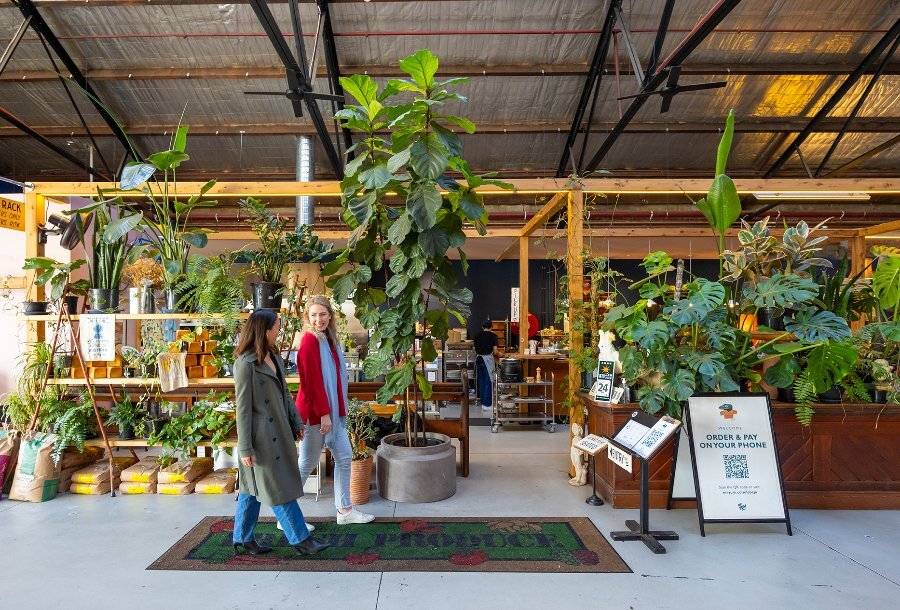 Two women walking into a cafe in a warehouse, with plant-filled shelves dividing the space.