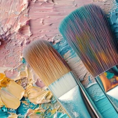 Artist paintbrushes resting diagonally over a colourful, textured canvas or palette with thick smears of pink, blue, yellow, and white paint.
