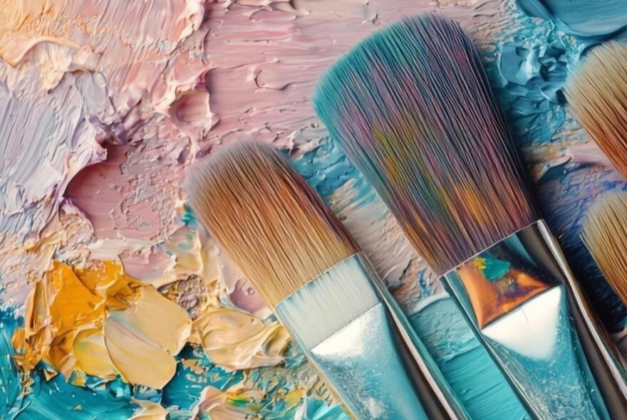 Artist paintbrushes resting diagonally over a colourful, textured canvas or palette with thick smears of pink, blue, yellow, and white paint.