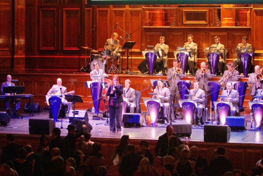 Big band performing seated on a purple-lit stage.