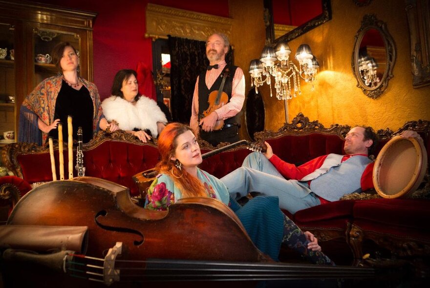 Performers reclining in a bohemian lounge setting with instruments, candelabra and mirrors.