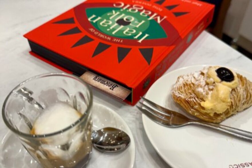 An empty coffee glass, pastry with fork on a plate and red-covered book on a table.