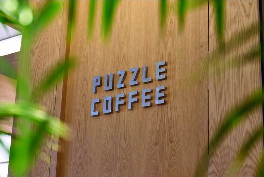 Puzzle Coffee sign on a wooden wall with ferns blurred in the foreground. 