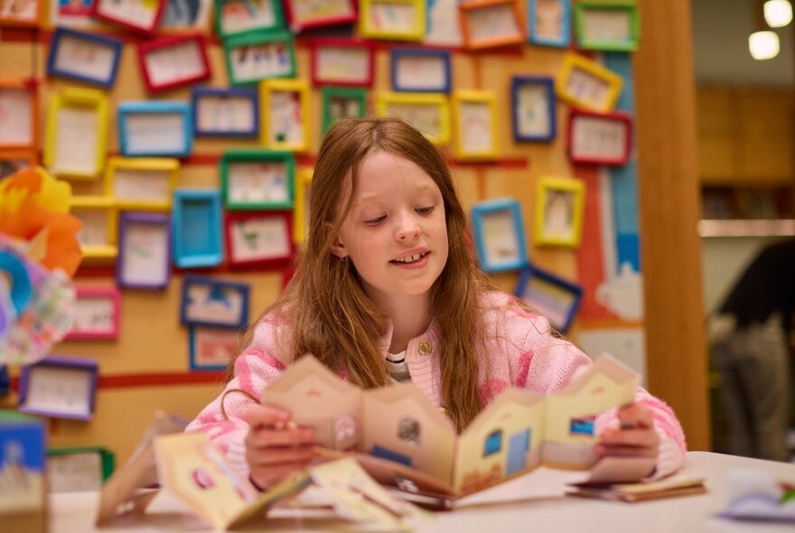 A primary-aged child sits at a table looking at a craft project in a room that features a colourful display of framed artwork or pictures hung in a grid pattern on the wall behind the table.