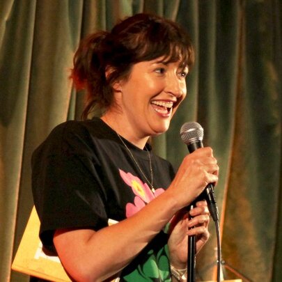 Comedian Celia Pacquola talking into a microphone she holds in her hand,  performing live on stage.