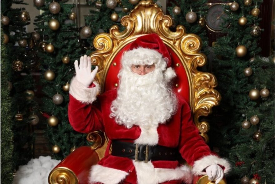 Santa with his hand raised seated in a large red and gold chair in front of a Christmas tree.