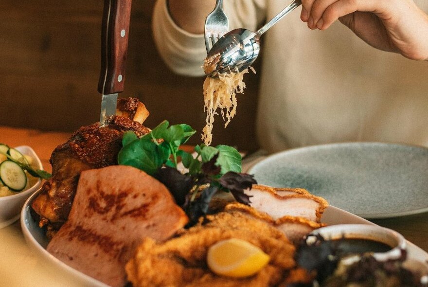 Hands using cutlery to select food from a large shared plate of thinly sliced cabbage, schnitzel, sausage, and other foods.