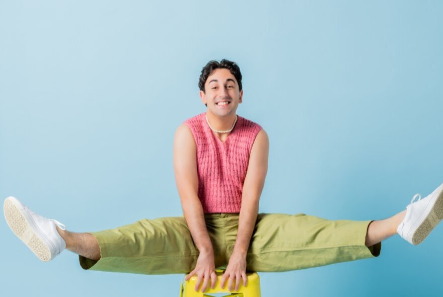 A man in a gymnastics horse pose with legs straight out, smiling happily against a blue background.