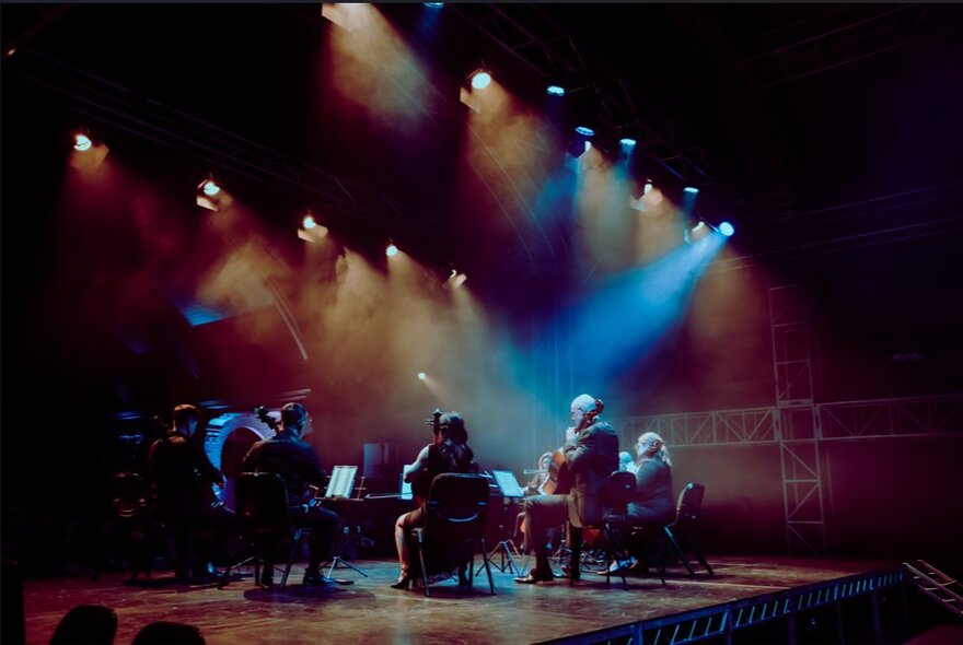 Seated cellists on a stage lit by turquoise spotlights.