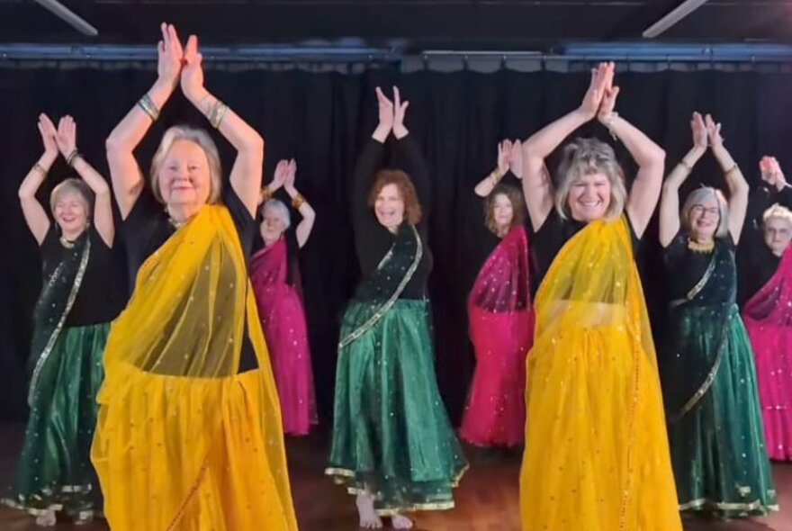 A group of older women dancing in colourful saris with their hands meeting above their heads. 