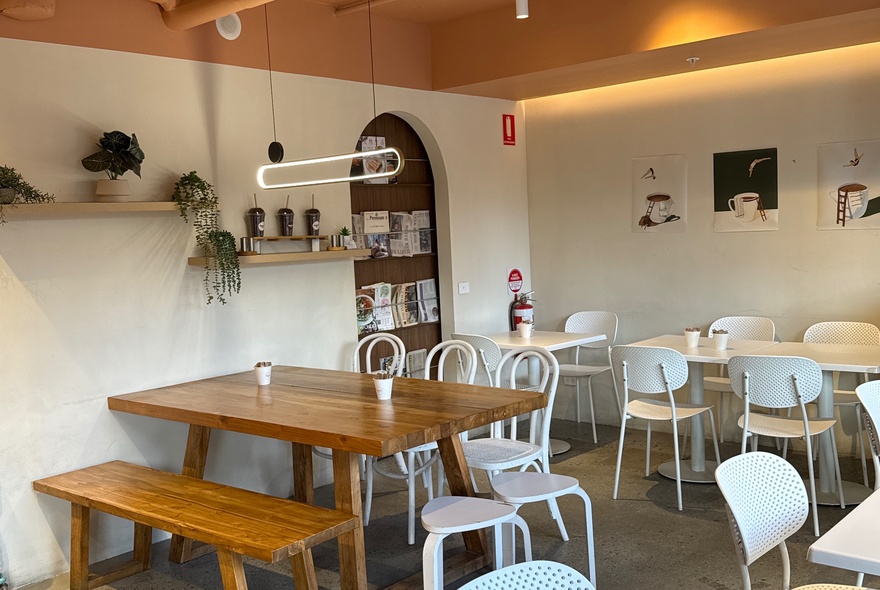 Cafe interior with wooden tables and white chairs in a white-painted space with plants on shelves.