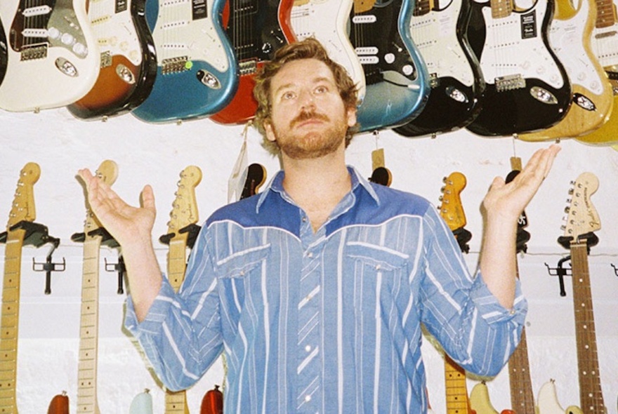 A man with his arms bent at the elbows and raised to shoulder height either side of him, standing in front of a wall of electric guitars.