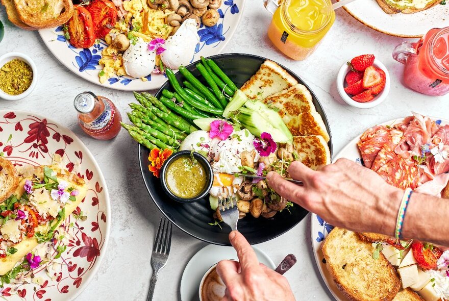 Hands cutting into a pan of grilled food with asparagus on a white table with other dishes of Italian food.