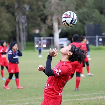 A young female soccer player heading the ball, wearing a red soccer uniform; teammates in the background.