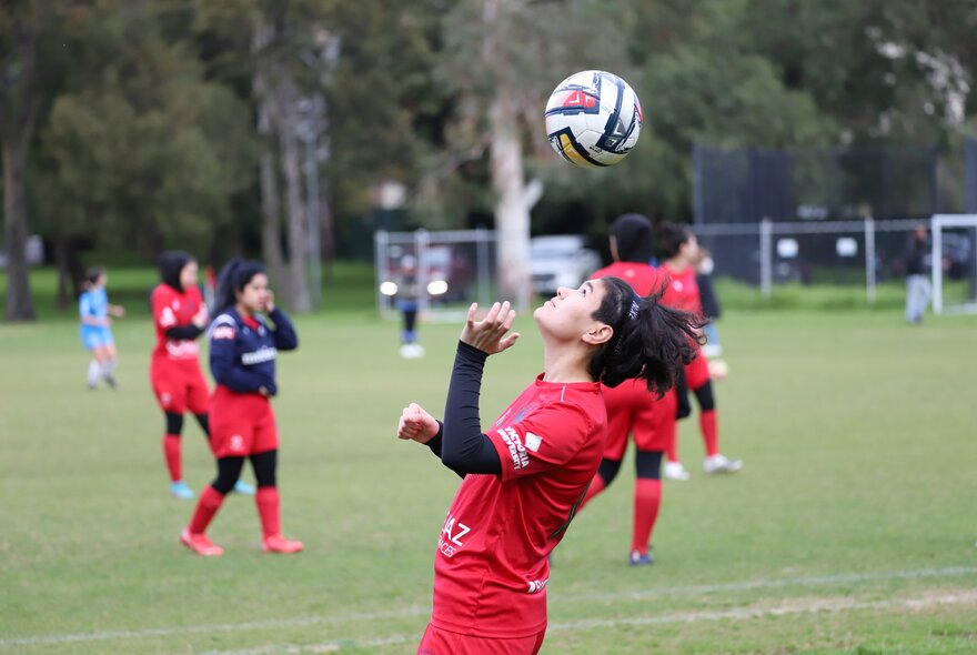 A young female soccer player heading the ball, wearing a red soccer uniform; teammates in the background.