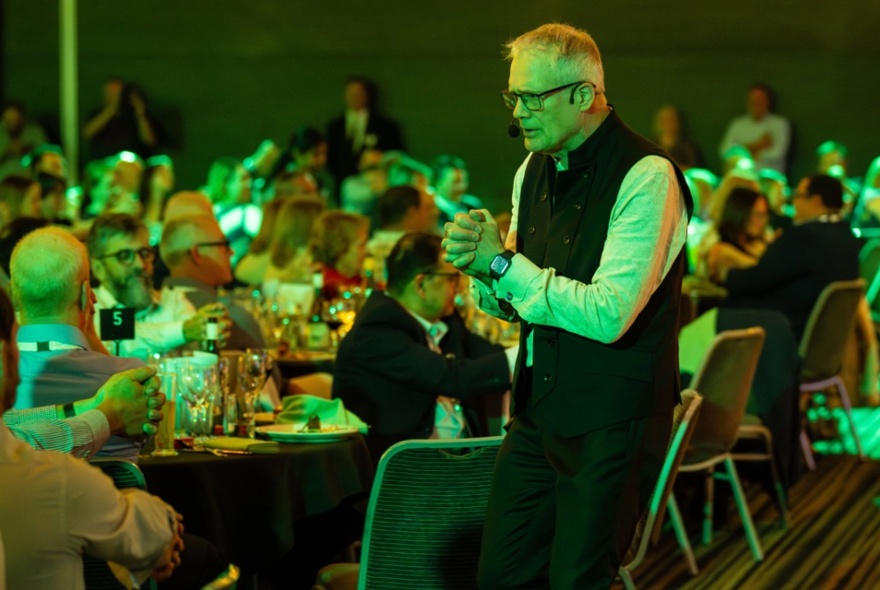 Entertainer wearing a dark waistcoat and glasses,  with hands clasped, performing to tables of seated diners.