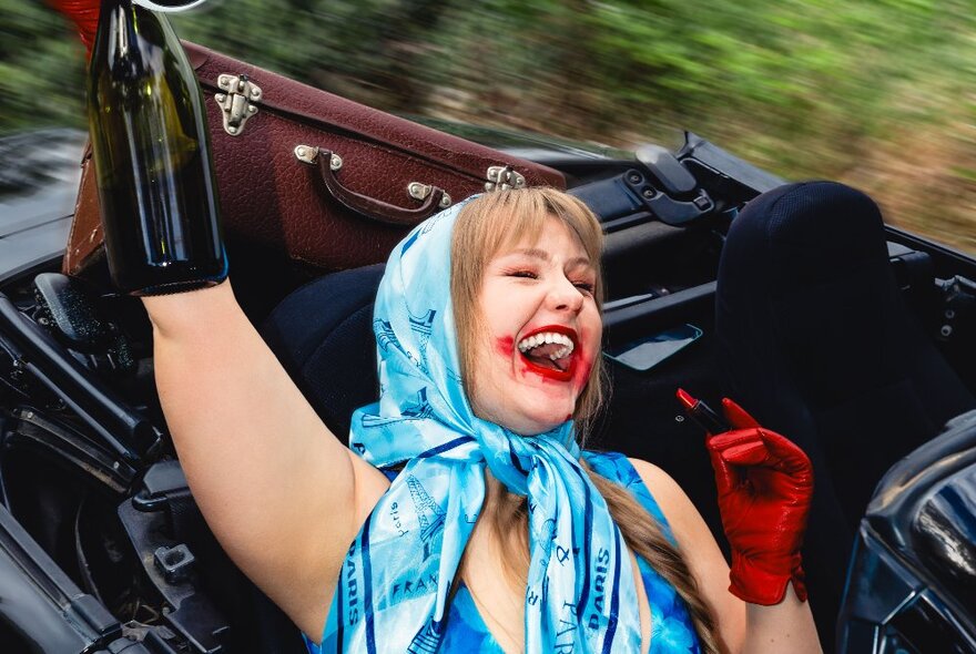 Comedian wearing headscarf and smudged lipstick, seated in an open-top car with her hand raised and laughing.