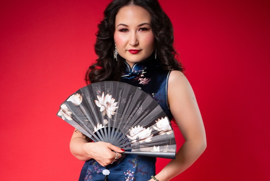 Comedian and cabaret artist Kelly Lim Harris holding an open fan against a red background.