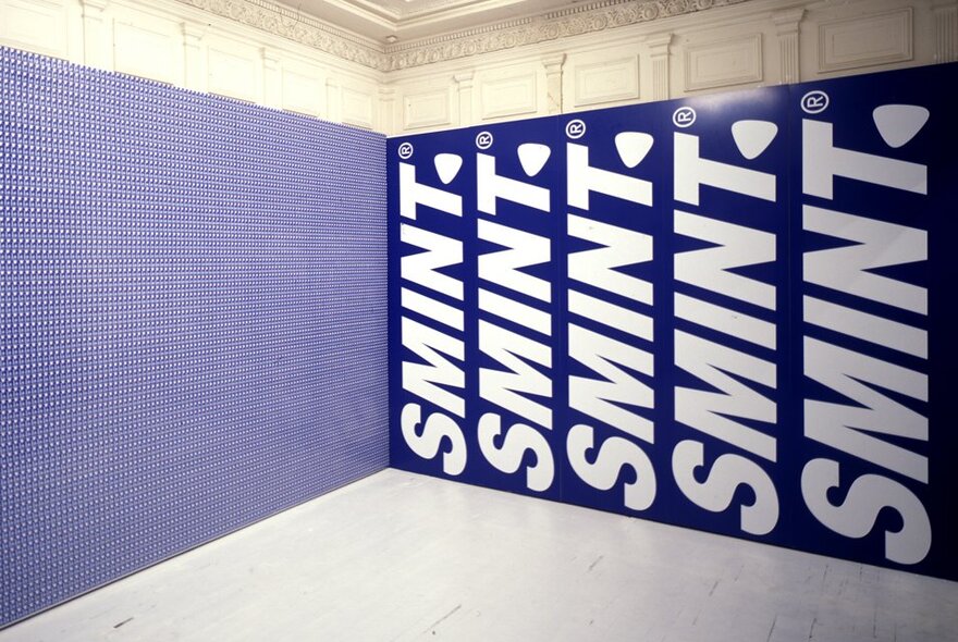 2000 Red Bull cans stacked against a wall in a gallery setting, with SMINT advertising signs lining the perpendicular wall.