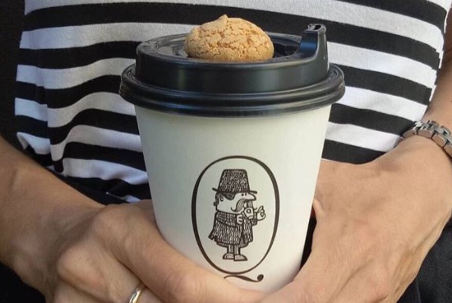 Hands holding a takeaway coffee cup with a small biscotti balanced on the lid of the cup.