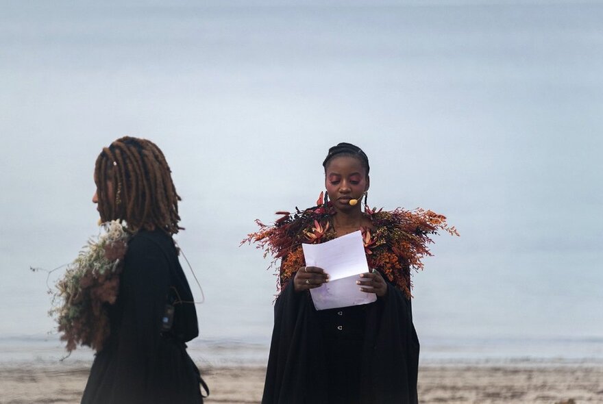 People wearing indigenous cloaks standing on a beach, one reading from a white sheet of paper.